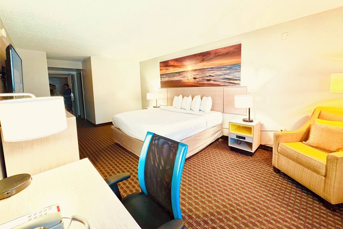 A hotel room with a bed, desk, chair, and a painting on the wall.