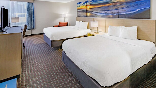 A hotel room with two beds, a desk, a TV, and a large painting of a sunset over the ocean.