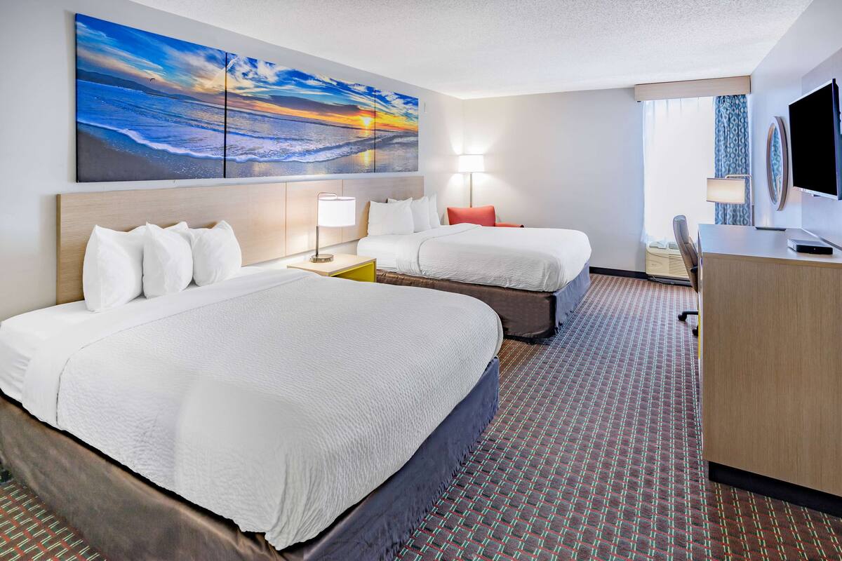 A hotel room with two beds, a desk, a TV, and a large painting of a sunset over the ocean.