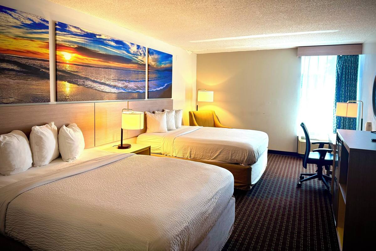 A hotel room with two beds, a desk, a chair, and a large window with curtains.