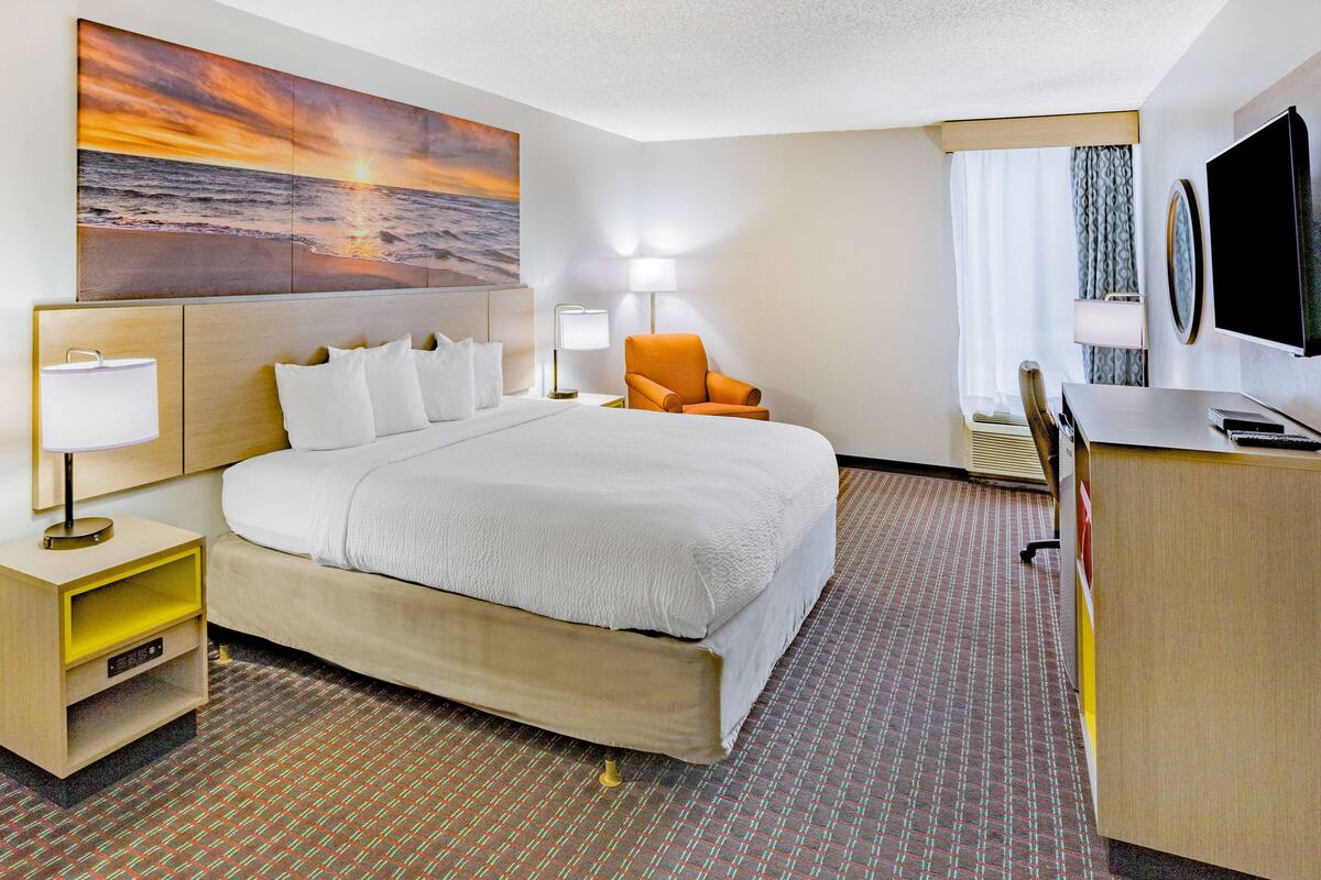 A hotel room with a large bed, a desk, a chair, a TV, and a painting of a sunset over the ocean.