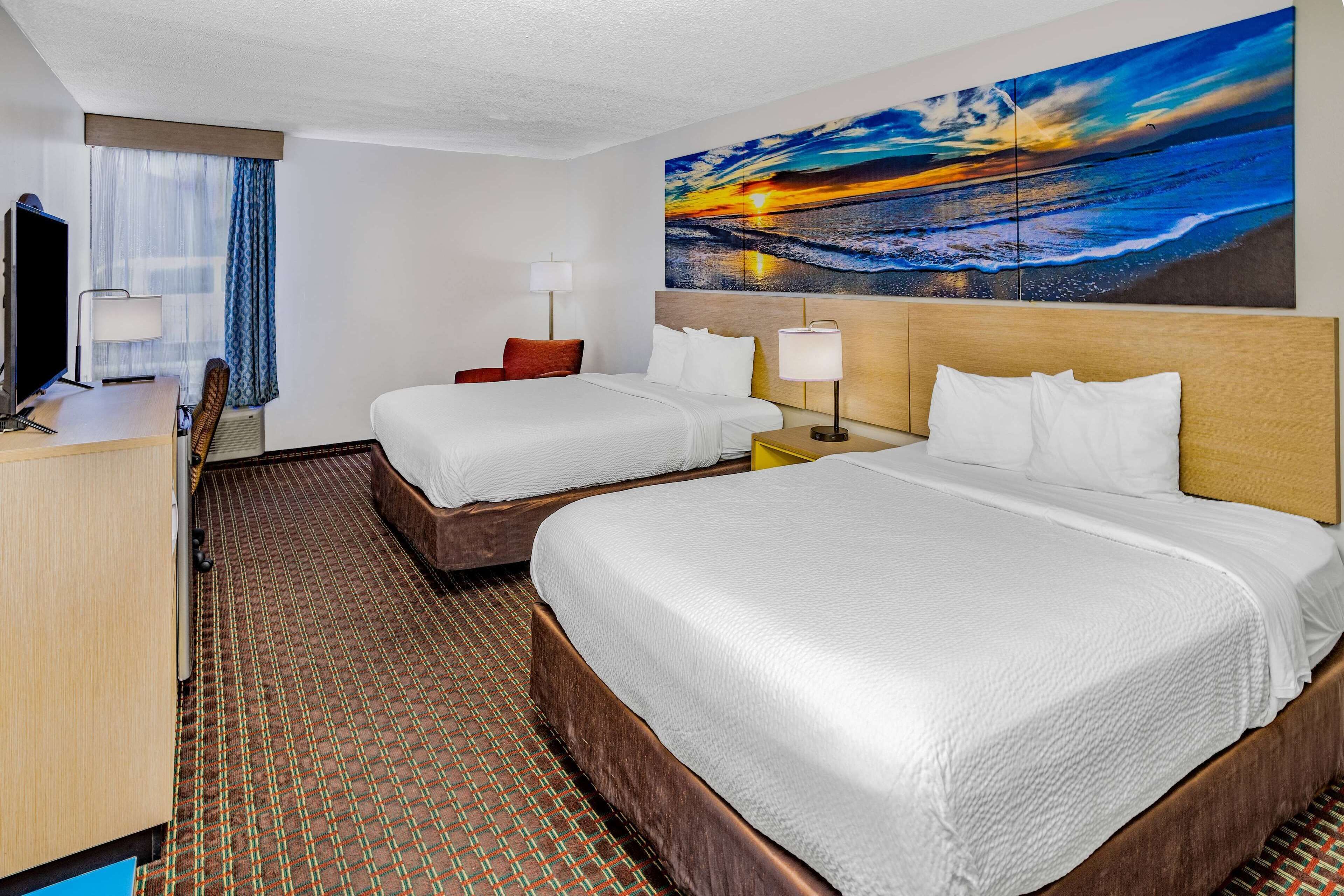 A hotel room with two beds, a desk, a chair, a TV, and a large painting of a sunset over the ocean.