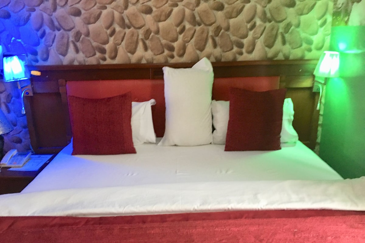 A bed with white bedding and red pillows, a stone wall behind, and a nightstand with a lamp.