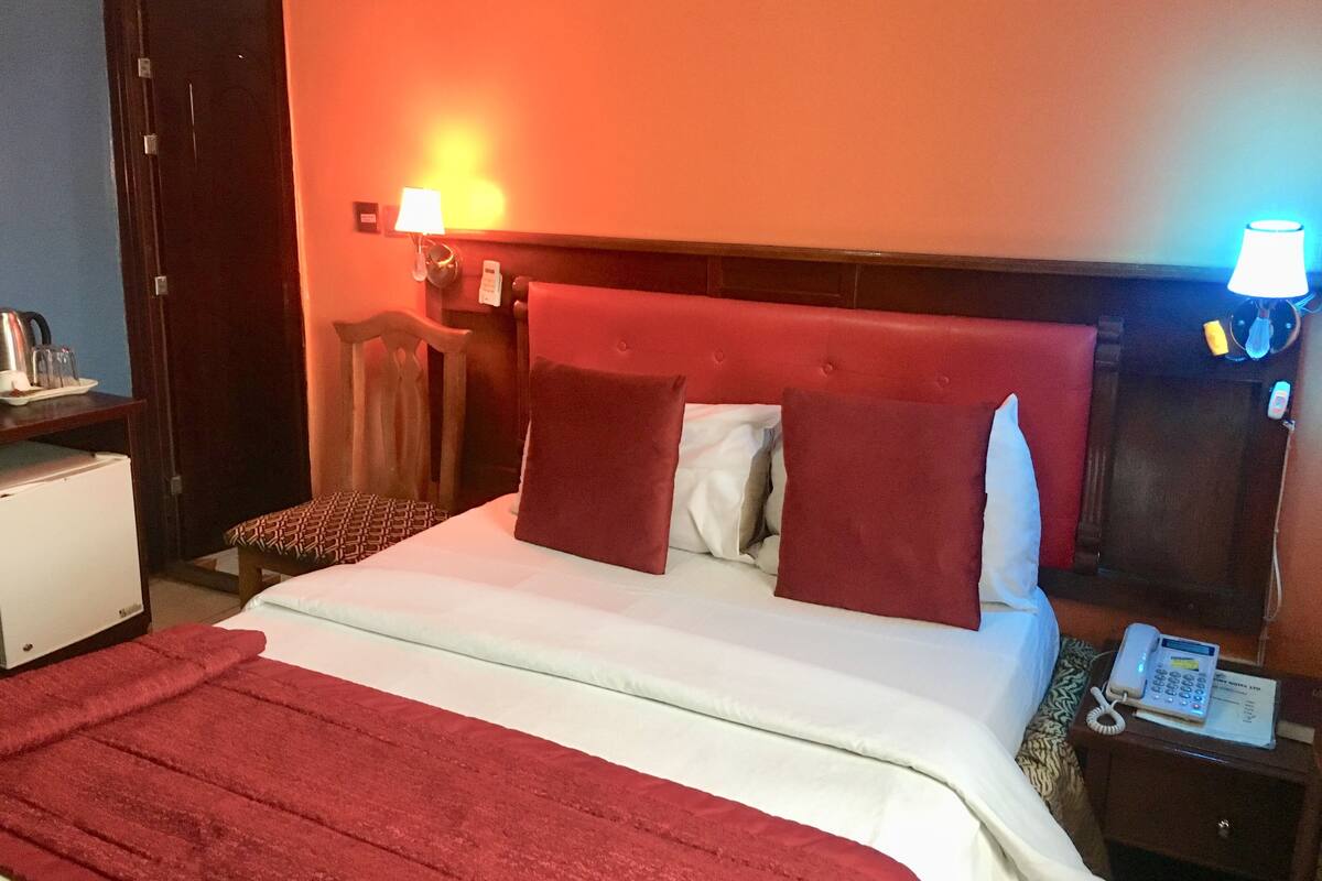 A hotel room with a bed, a nightstand with a phone, a desk with a lamp, and a chair.