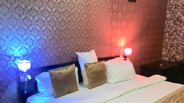 A neatly made bed with white sheets and multiple pillows, including a white and a brown one. There are two bedside lamps with red and blue lighting. - Definite Destiny Hotel (Lagos)