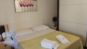 A hotel room with a bed, white pillows, a yellow bedspread, folded towels on the bed, a nightstand with a lamp, and a painting of pink flowers on the wall. - Al Torrione (Reggio Calabria)