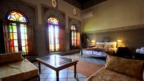 Quarto com cama, mesa e dois sofás. Três grandes janelas com vitrais.