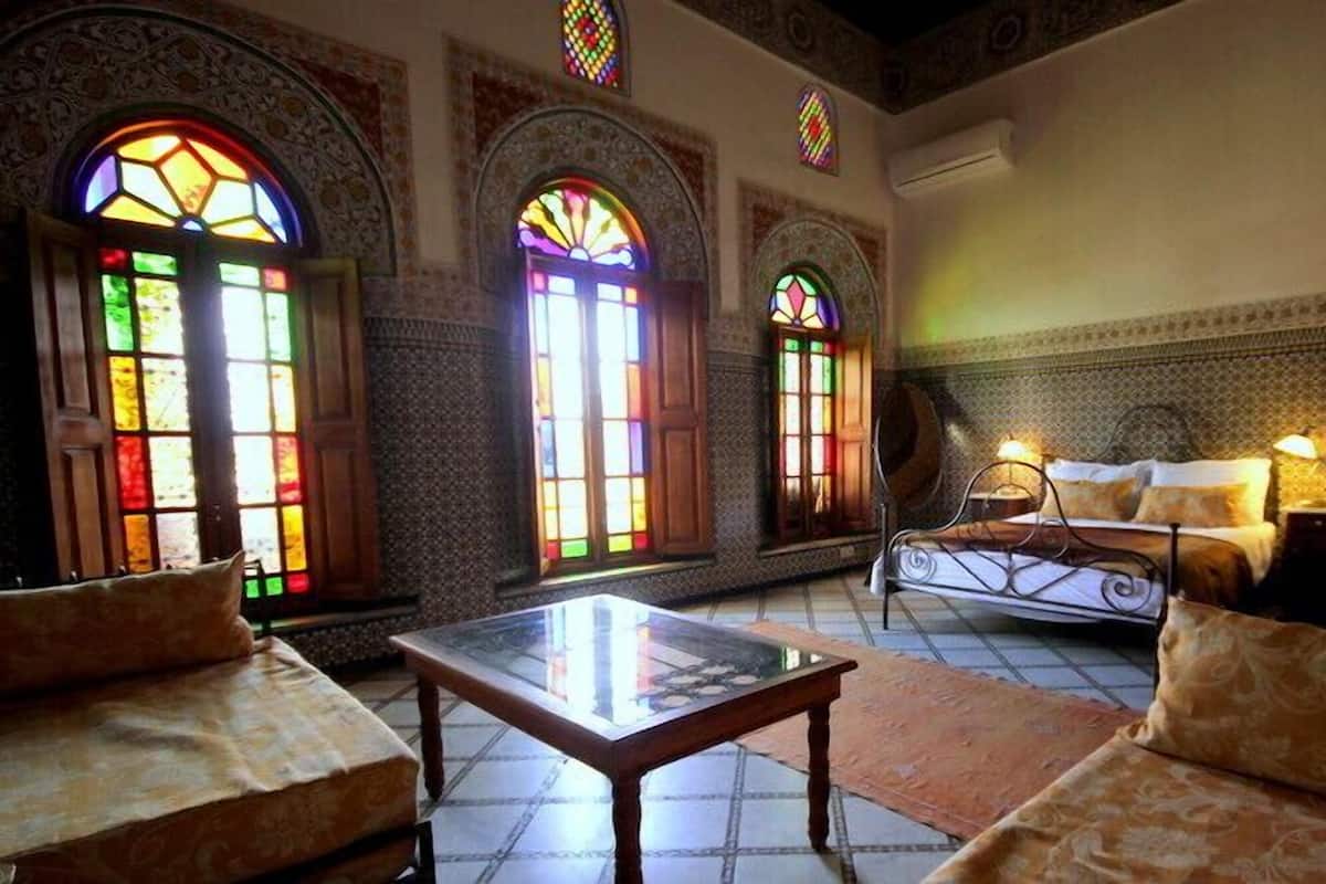A room with a bed, a table, and two sofas. There are three large windows with stained glass.
