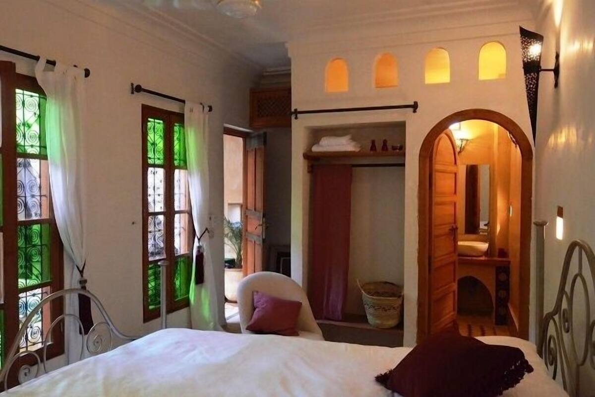 A bedroom with a bed, a chair, a wardrobe, and a bathroom visible through an archway.