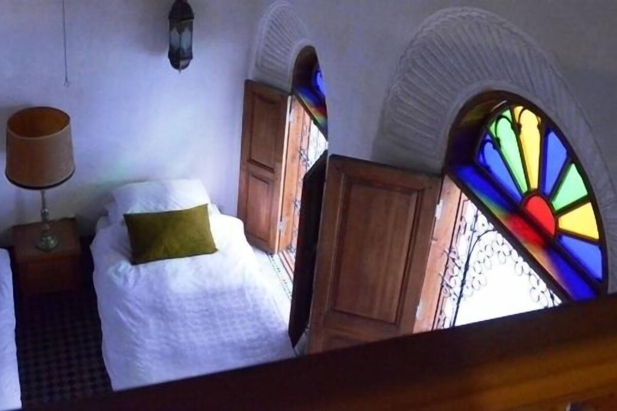A room with two beds, a lamp, a wooden cabinet, and a stained glass window.