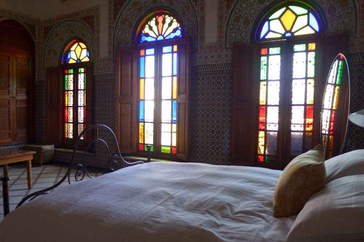 A room with a bed, a bench, and three arched windows with colorful stained glass.