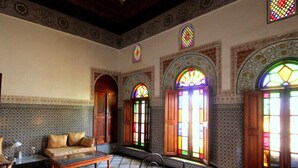 Quarto tradicional com cama, sofá e mesa. Vários janelões com vitrais.