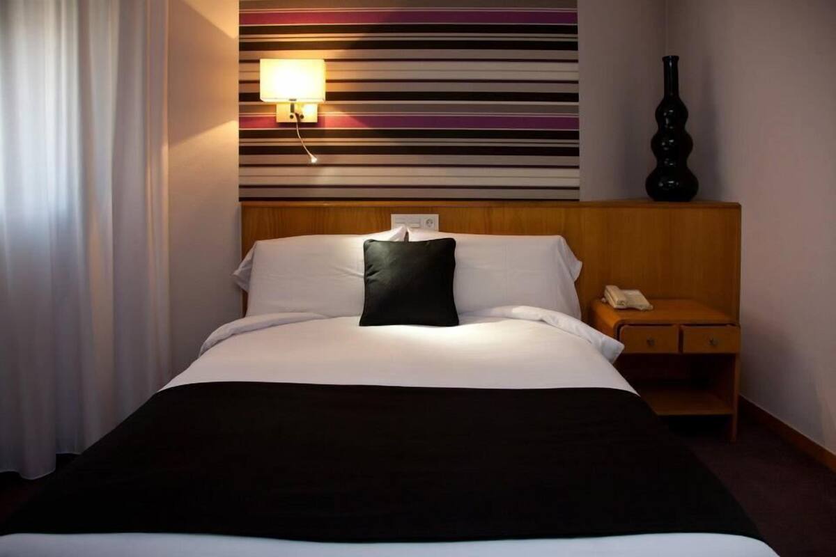 Una habitación de hotel con una cama, una mesita de noche, una lámpara y un jarrón.