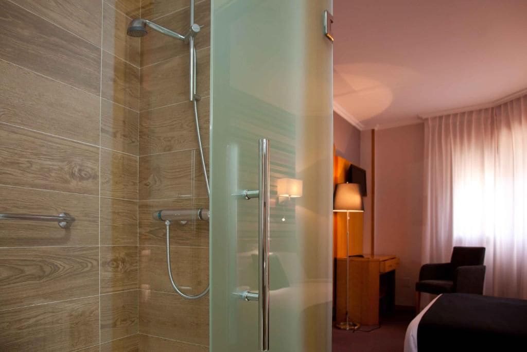 Un baño moderno con cabina de ducha de vidrio, suelo de madera, mesita de noche con lámpara y una cama.