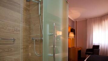 Un baño moderno con cabina de ducha de vidrio, suelo de madera, mesita de noche con lámpara y una cama.