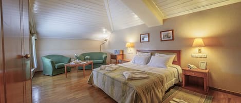 Ett hotellrum med ett träpanelat tak, en säng med ett beige överkast, en grön fåtölj, ett litet bord med en vas och ett nattduksbord med en lampa.