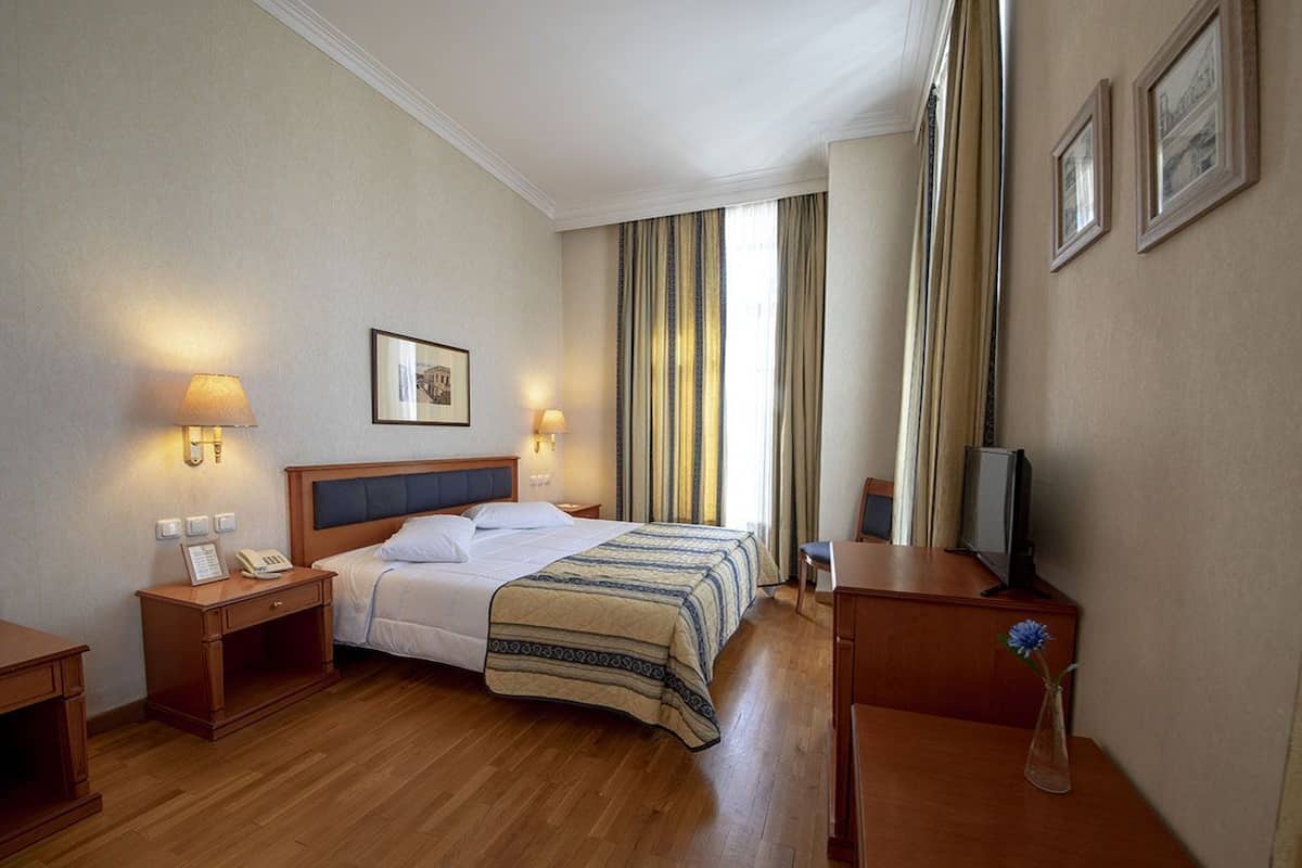 A hotel room with a bed, bedside lamps, a desk, a chair, a TV, and a vase with a flower.