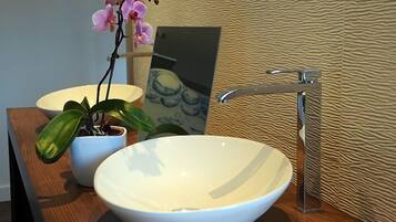 Une salle de bain avec un lavabo blanc en forme de cuvette, une orchidée en pot et un miroir posé sur un comptoir en bois.