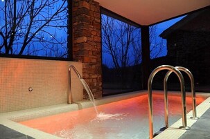 Una bañera de hidromasaje con un pilar de piedra y barandillas metálicas, frente a un fondo de árboles y un edificio.