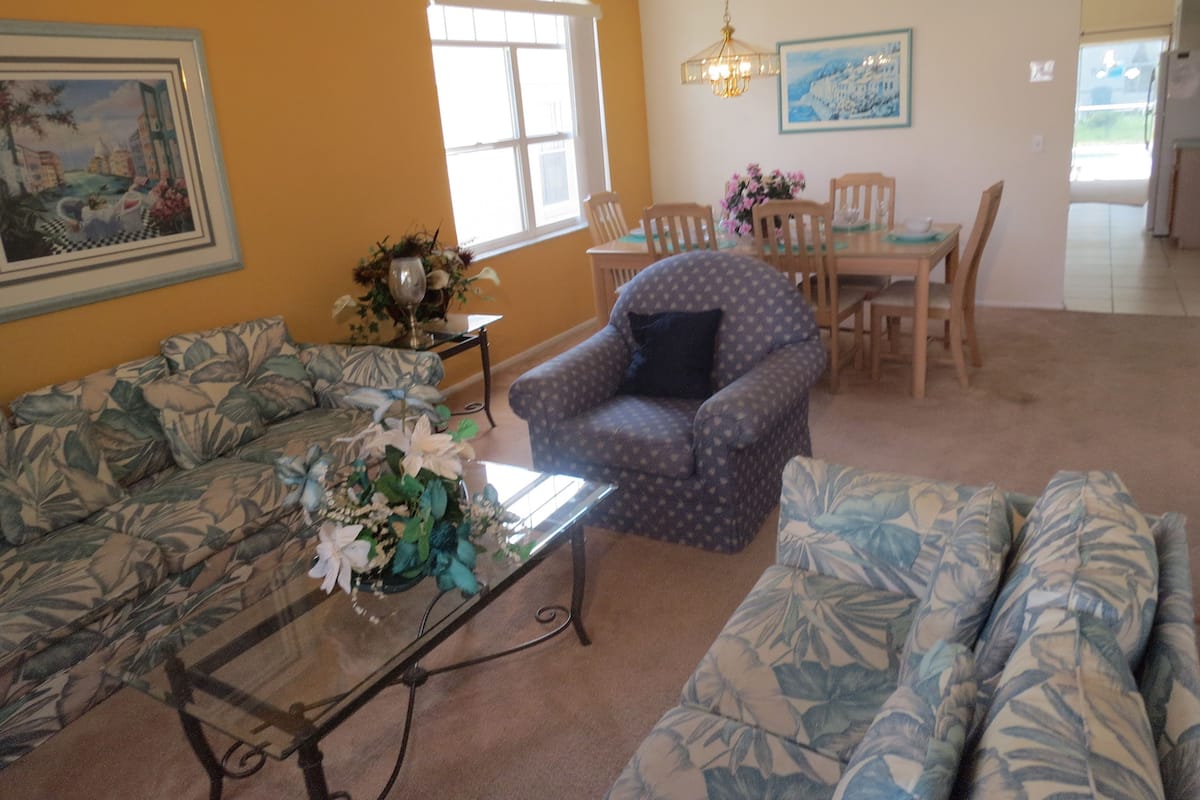 Uma sala de estar com sofá, poltrona e mesa de centro. Há uma mesa de jantar com cadeiras e um lustre.