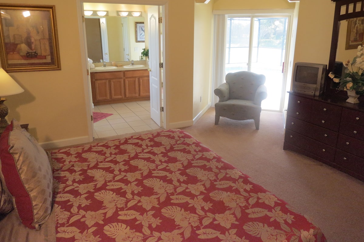 Quarto com carpete floral, uma cadeira, cômoda e banheiro visível por uma porta aberta.