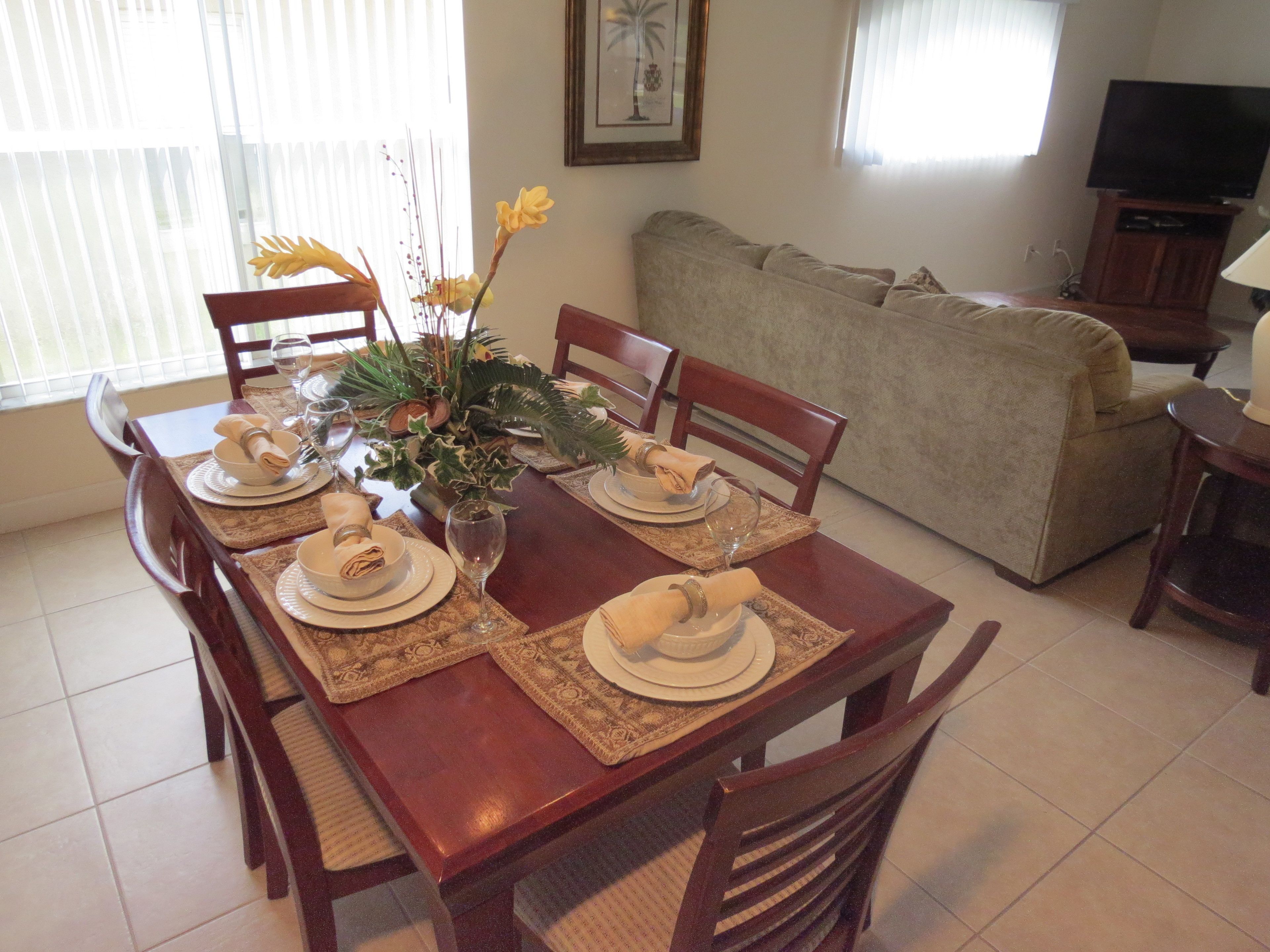 Un comedor con una mesa de madera dispuesta para cuatro personas, rodeada de sillas. Se ve un sofá y un televisor al fondo.