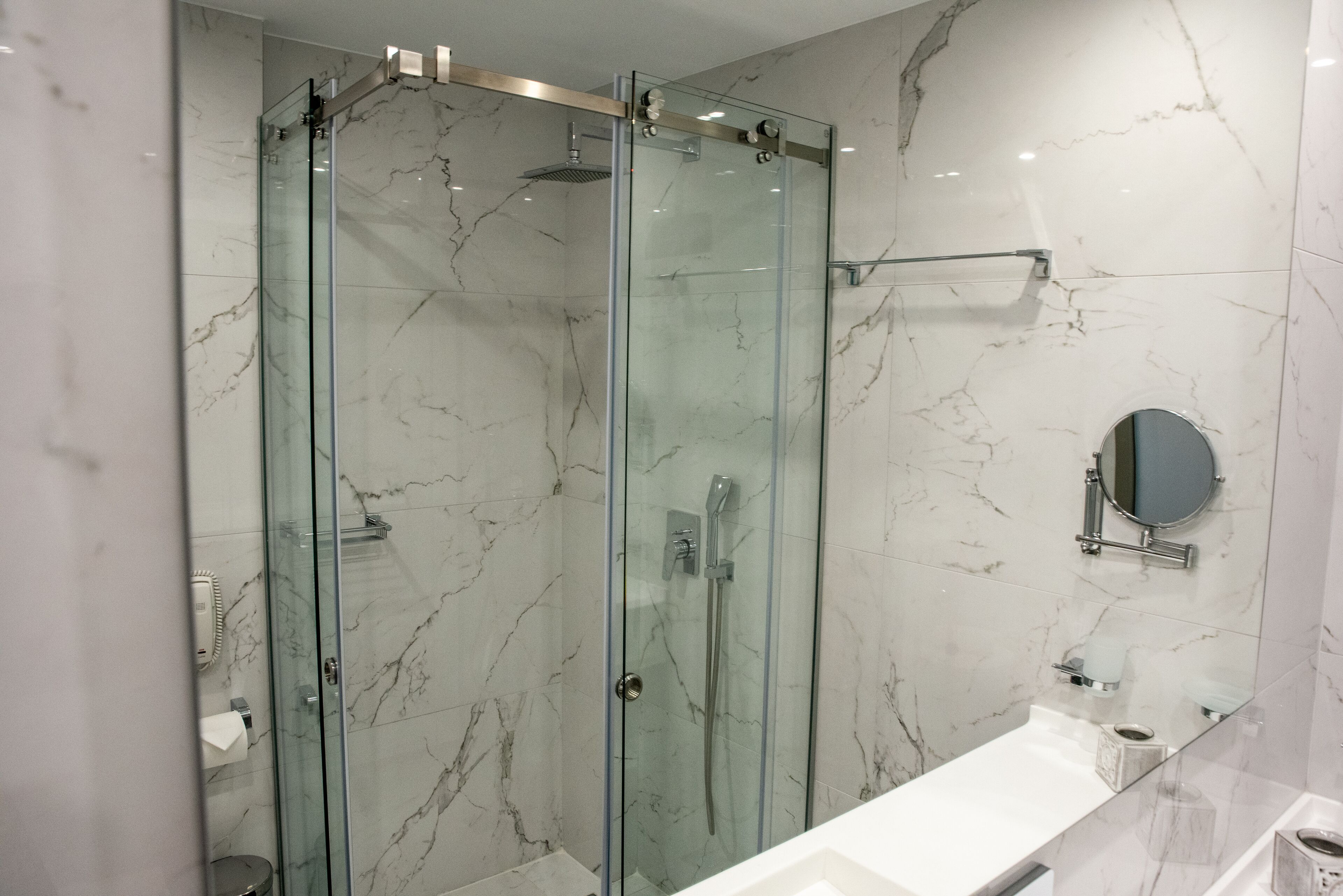 A modern bathroom with a glass-enclosed shower, marble walls, a white sink, and a round mirror.