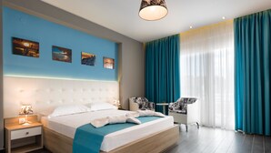 A modern hotel room with a large bed, a bedside table with a lamp, a chair, and a view of the city through sheer curtains. - Lagaria Hotel (Kassandra)