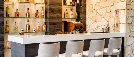 Zona de bar con mostrador blanco, tres taburetes y estantes llenos de botellas.