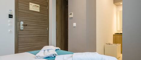 Habitación de hotel con puerta de madera, cama con cubrecama azul y pared blanca con termostato y pulsador de luz.