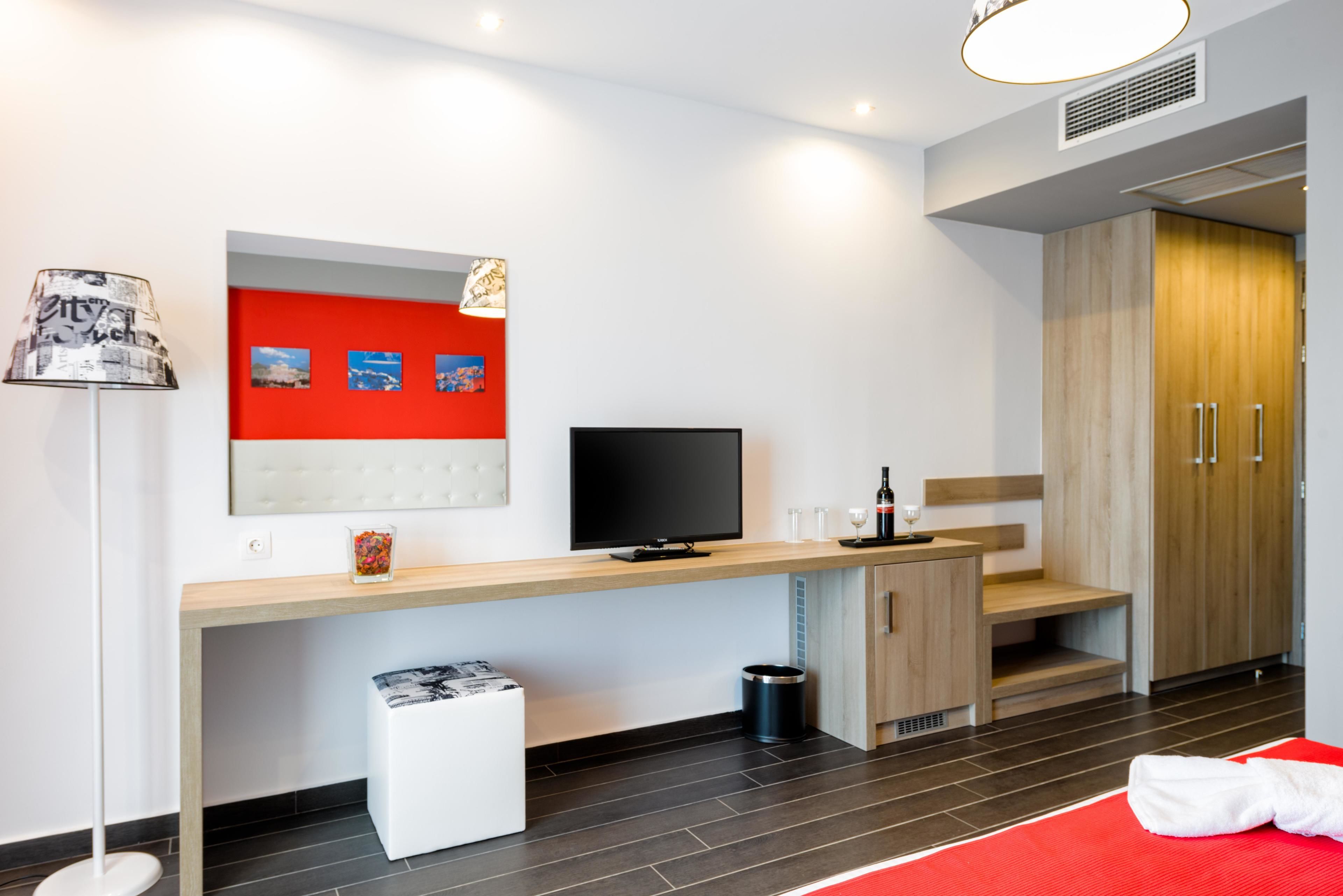 A modern hotel room with a wooden desk, a flat-screen TV, a red accent wall, a white ottoman, and a red mat.
