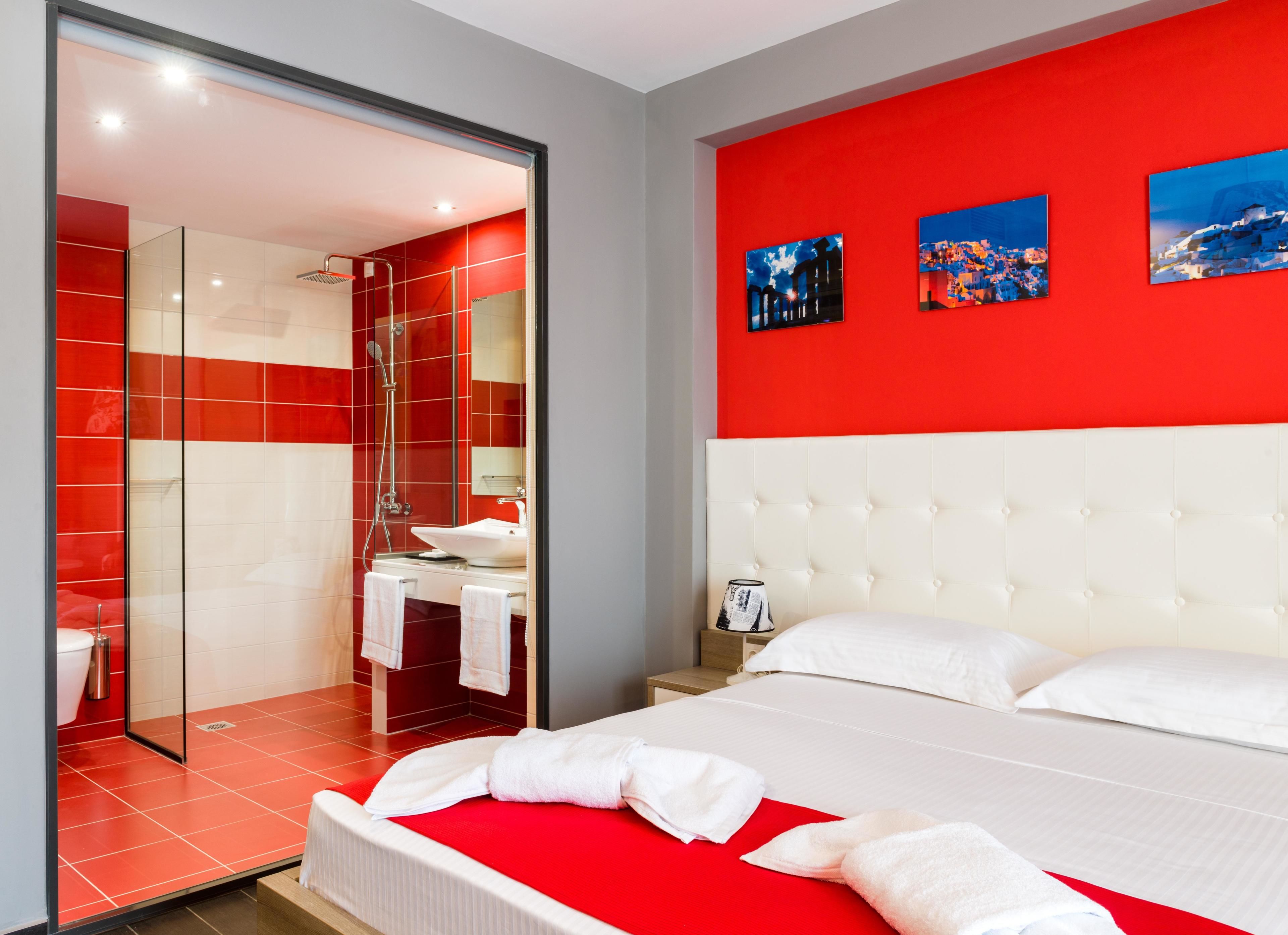 A modern hotel room with a red and white color scheme, a large bed, a bathroom with a glass shower enclosure, and artwork on the wall.