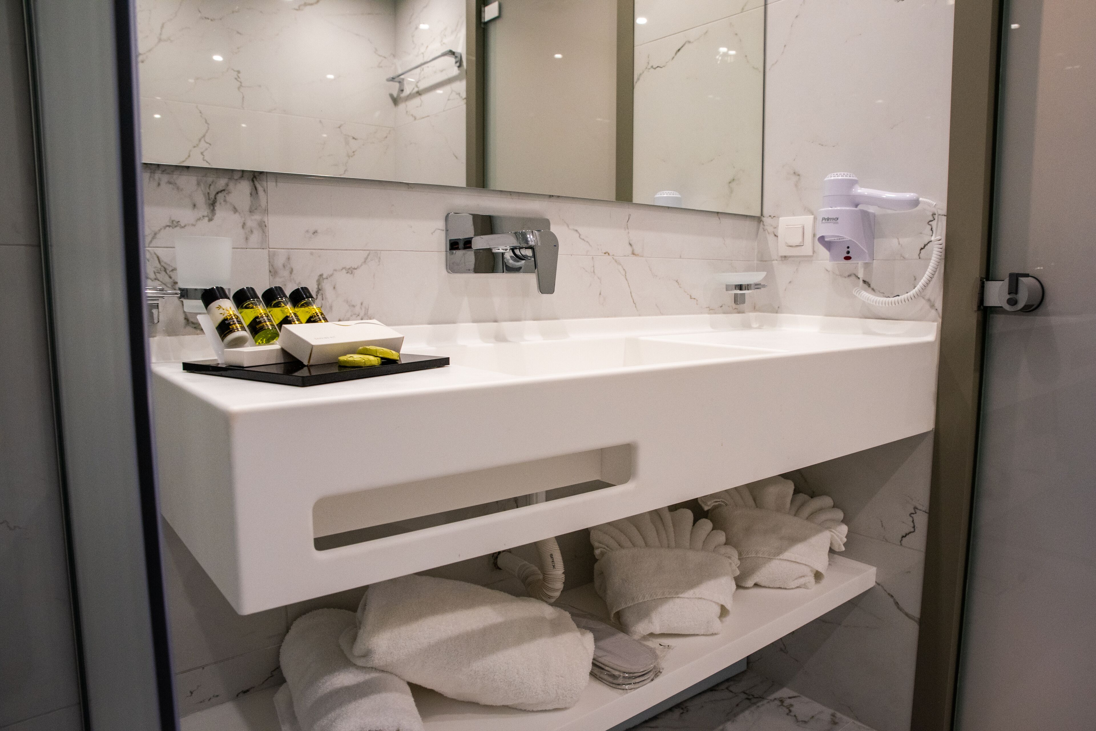 Une salle de bain moderne avec un lavabo blanc, un miroir, un sèche-cheveux et des serviettes soigneusement pliées.