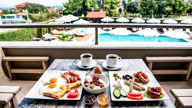 A breakfast table with plates of food, two cups of coffee, and a view of a pool and umbrellas. - Lagaria Hotel (Kassandra)