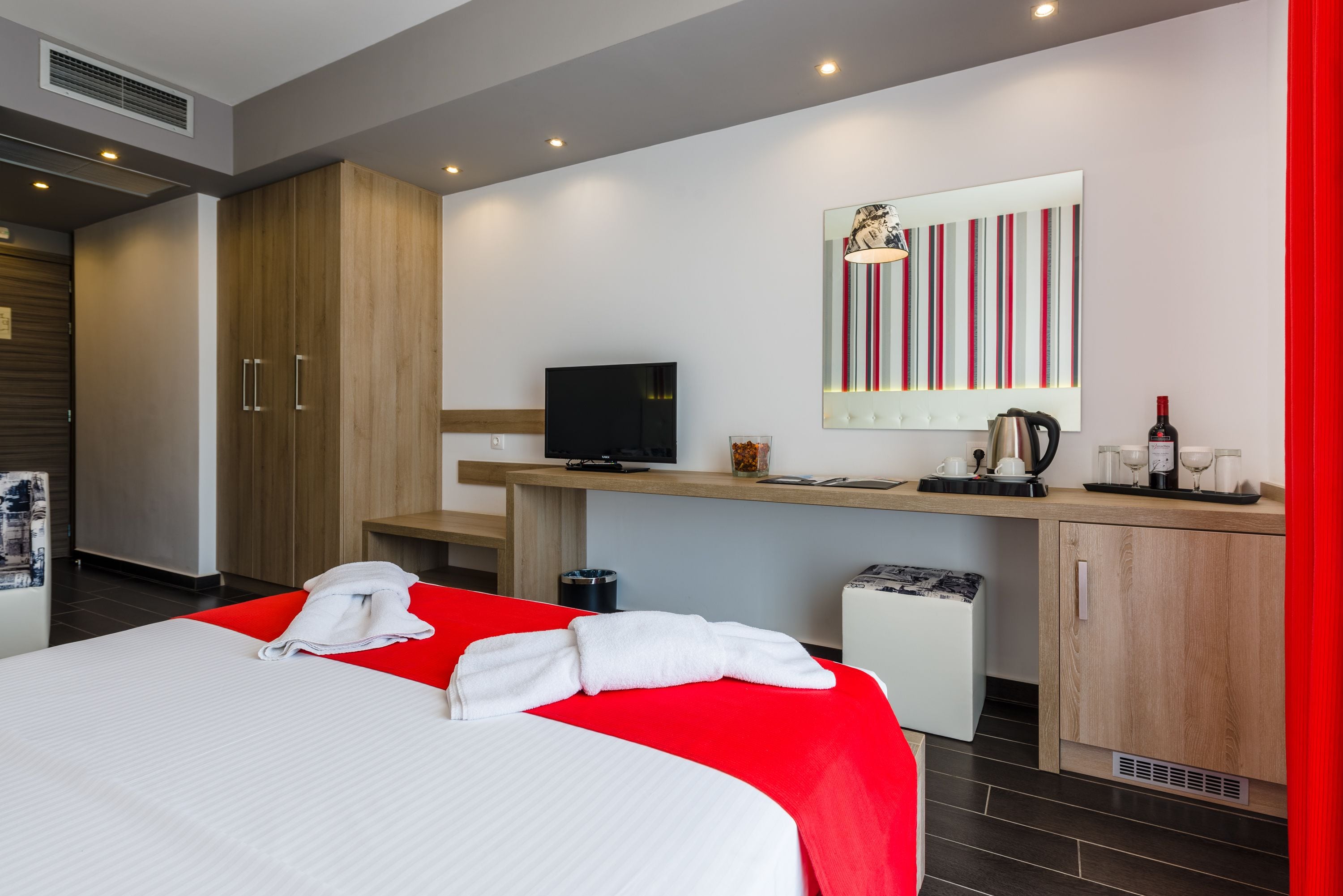 A hotel room with a bed, a desk with a television, a mirror, and a small kitchenette.
