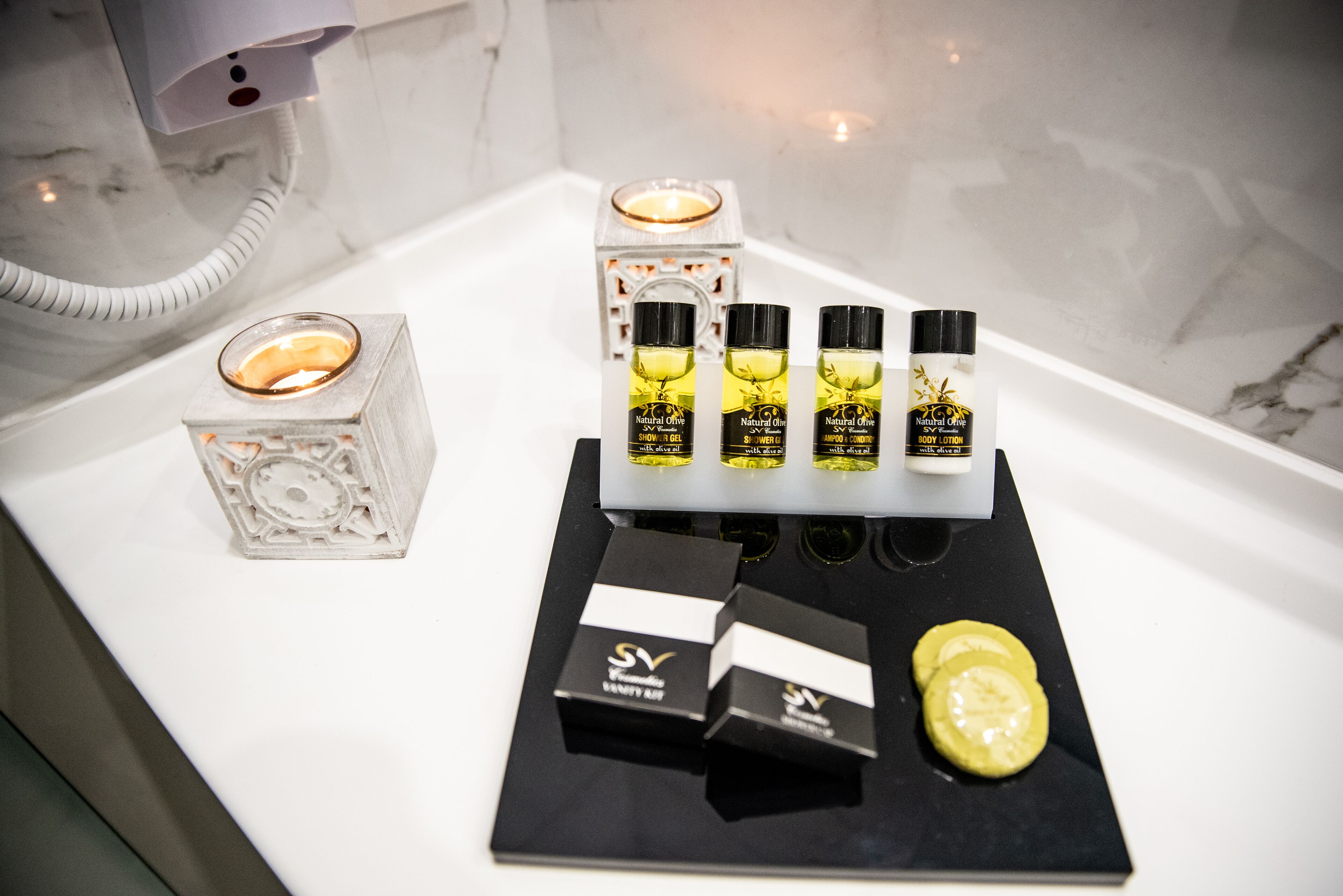 Un meuble-lavabo de salle de bain avec un plan de toilette en marbre blanc, un porte-bougie en verre et un ensemble de flacons de lotion pour le corps à base d’huile d’olive naturelle.