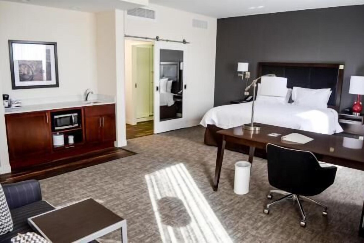 A hotel room with a bed, desk, chair, and a small kitchenette.
