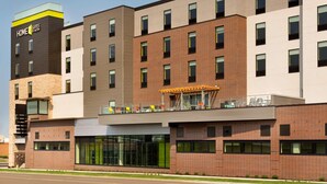 Een hotel met meerdere verdiepingen, modern van ontwerp, met een opvallend 'Home 2 Suites by Hilton' bord aan de linkerkant. Het gebouw heeft een combinatie van bakstenen en glazen gevels, en een dakterras met terrasmeubilair.