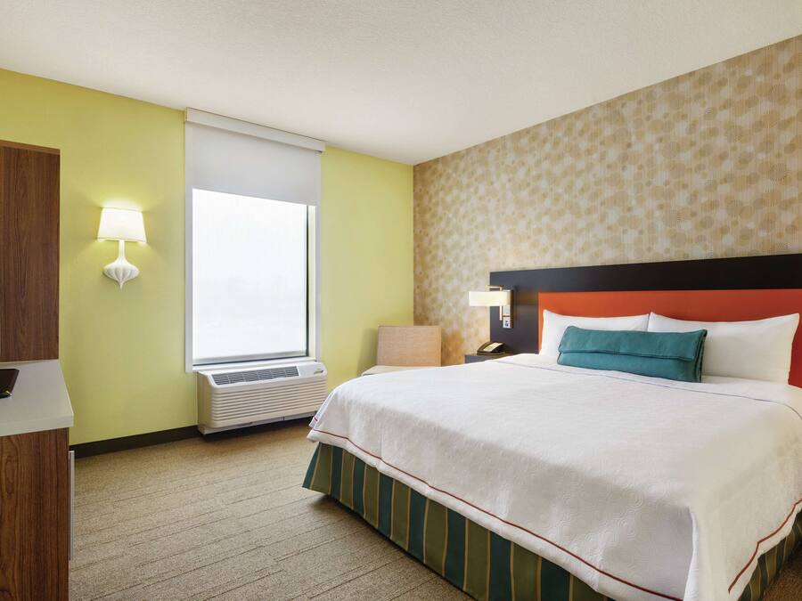 A hotel room with a bed, a TV, a lamp, a window, and a wall heater.