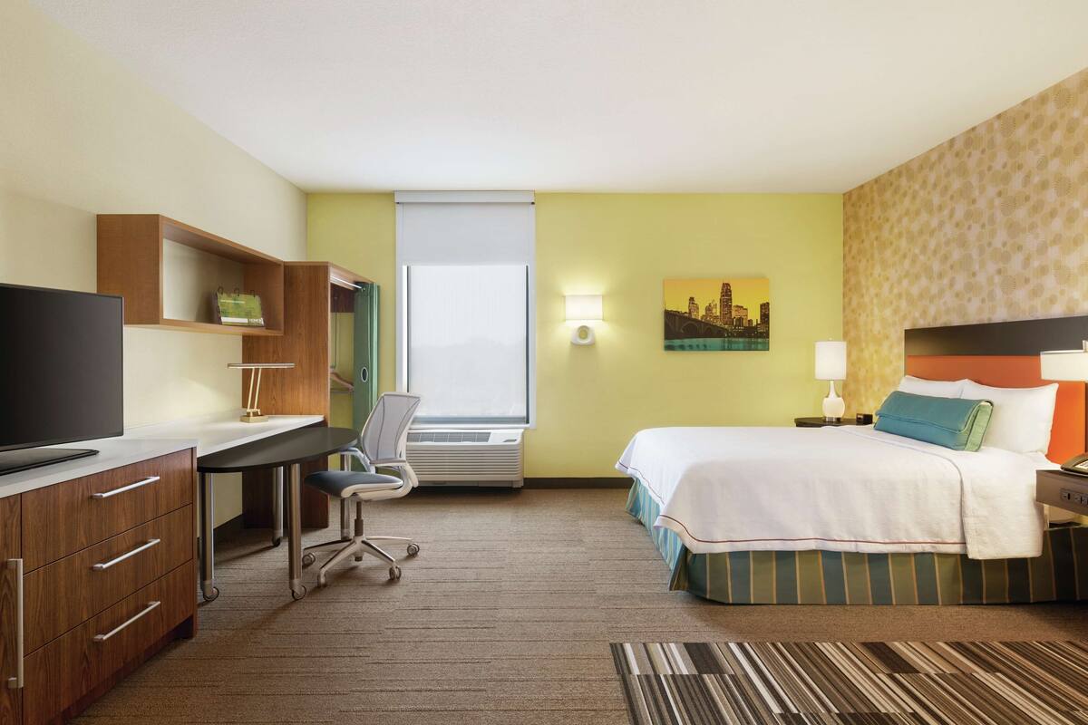 Une chambre d’hôtel équipée d’un bureau, d’une chaise, d’un lit et ornée d’un tableau représentant un paysage urbain.