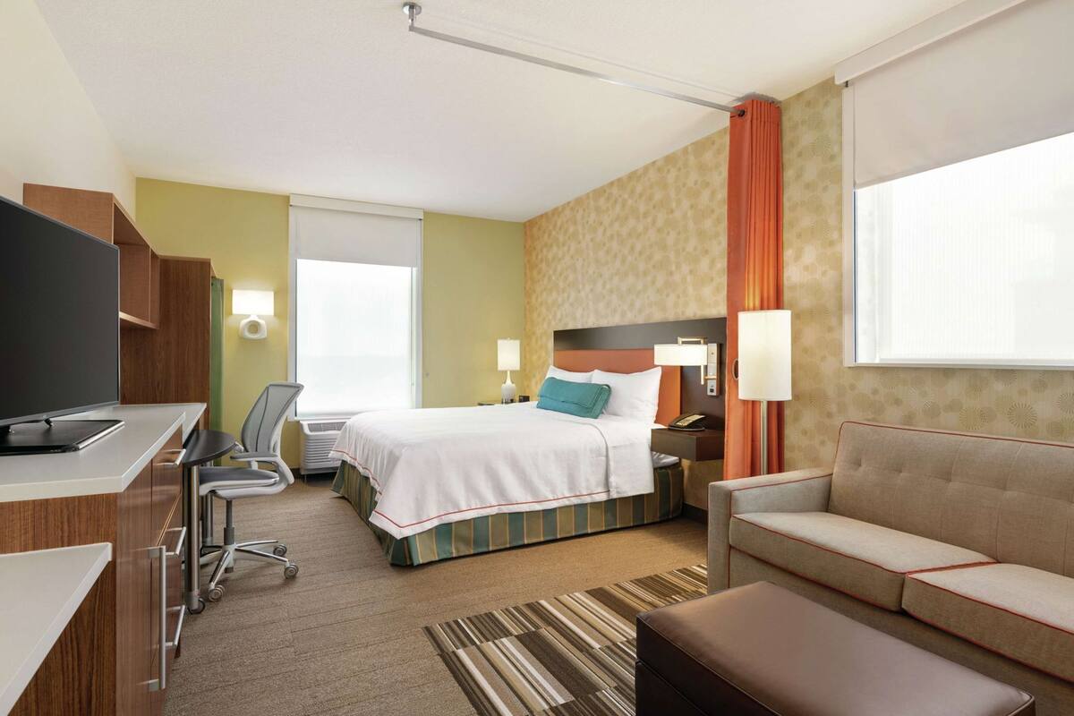 Quarto de hotel com cama, escrivaninha com computador, cadeira, sofá e televisão.