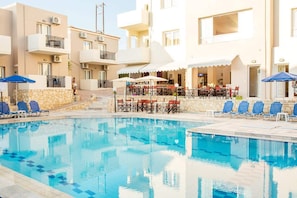 Une piscine entourée de bâtiments à plusieurs étages dotés de balcons et d’espaces de détente en plein air, avec des chaises longues et des parasols à proximité.