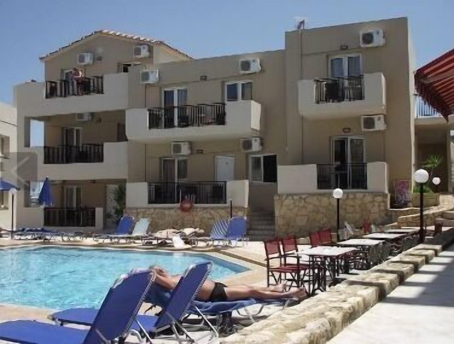 A poolside area with lounge chairs, a pool, and a building with balconies and air conditioning units.