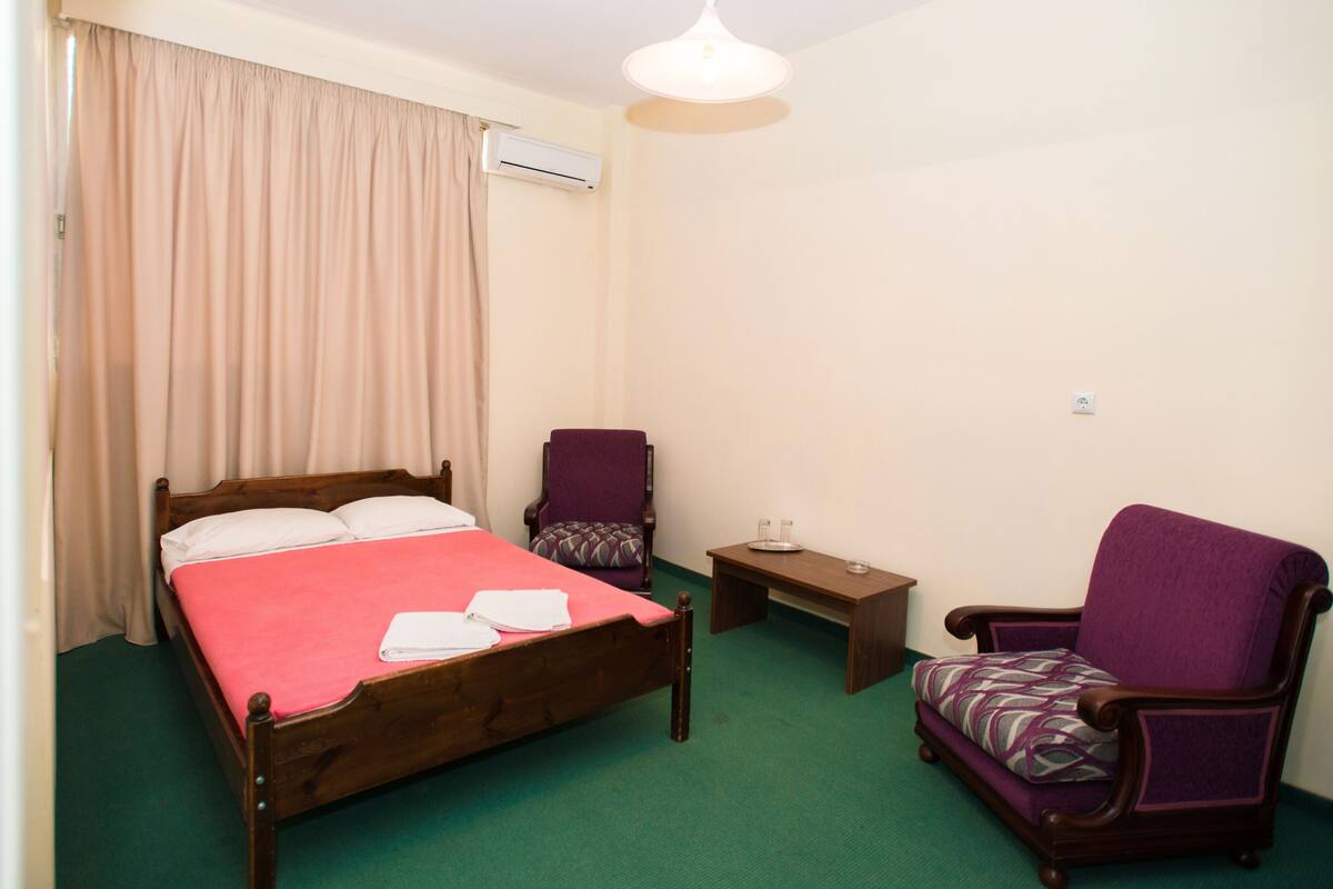A neatly arranged bedroom with a wooden bed, a purple armchair, a small bedside table, and a green carpet.