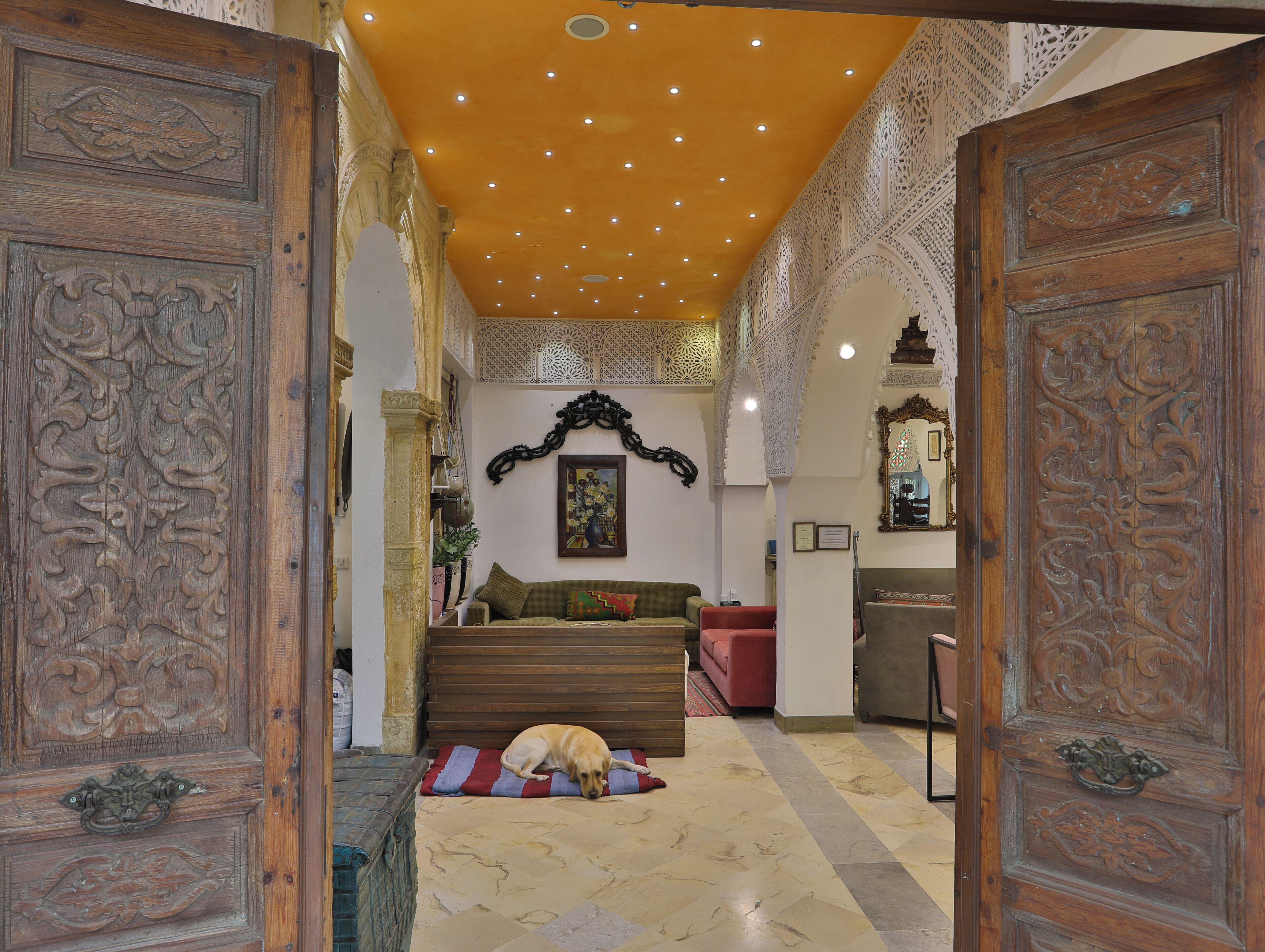 A hallway with ornate wooden doors, a dog resting on a mat, and a staircase leading to a room with a red sofa and a painting on the wall.