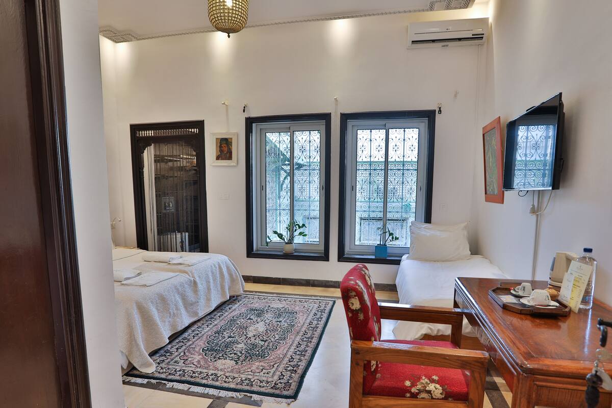Una habitación de hotel con una cama, un escritorio, una silla, un televisor y dos ventanas con rejas decorativas de hierro.