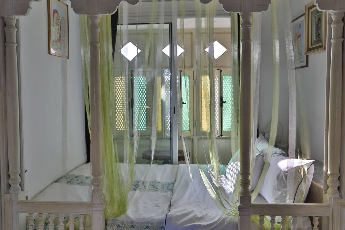 Una habitación con una cama bajo un dosel, un ventanal con diseño de rombos y una puerta con espejo.