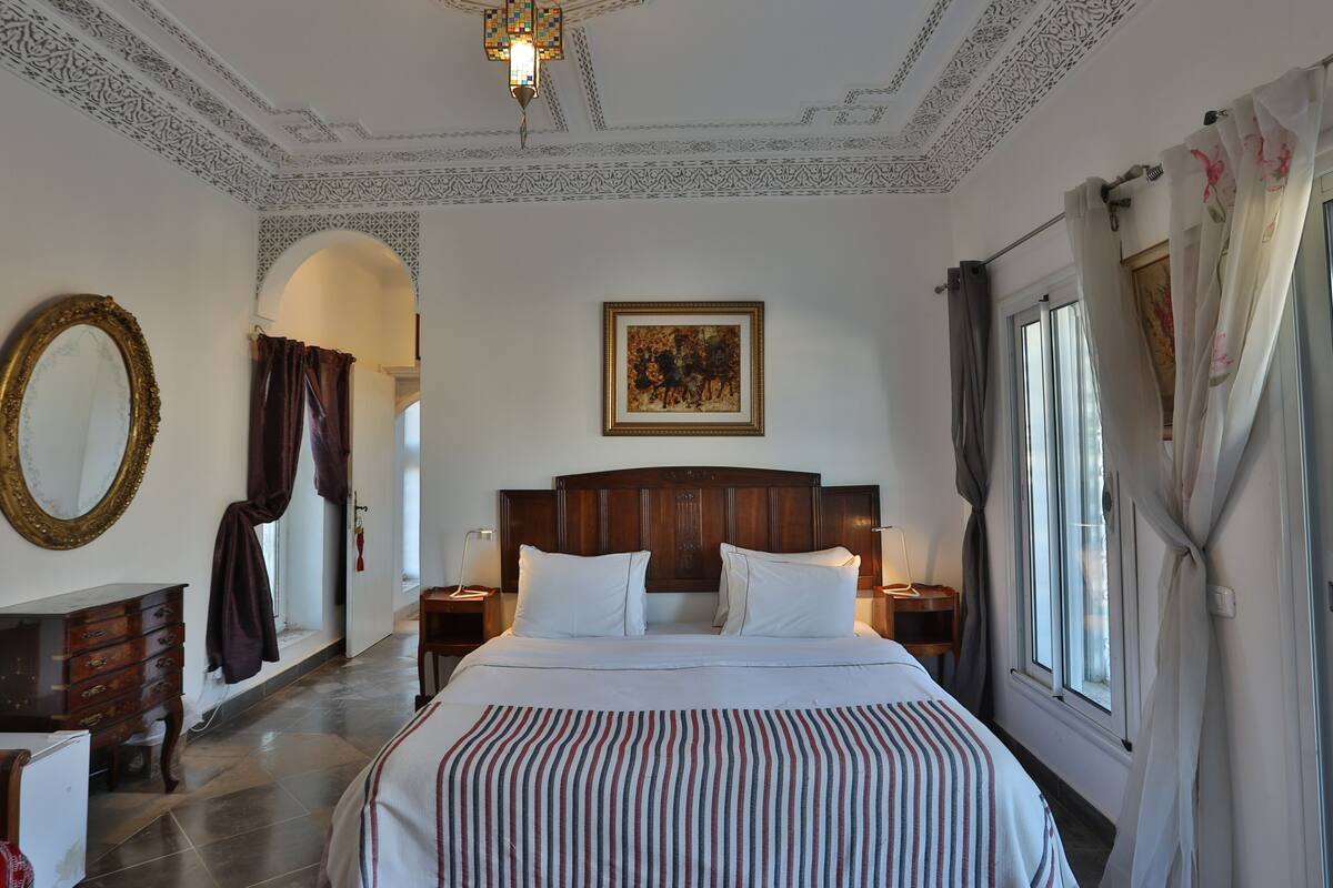 Una habitación de hotel con una cama grande, cabecera de madera, una araña, un cuadro en la pared y una ventana con cortinas.
