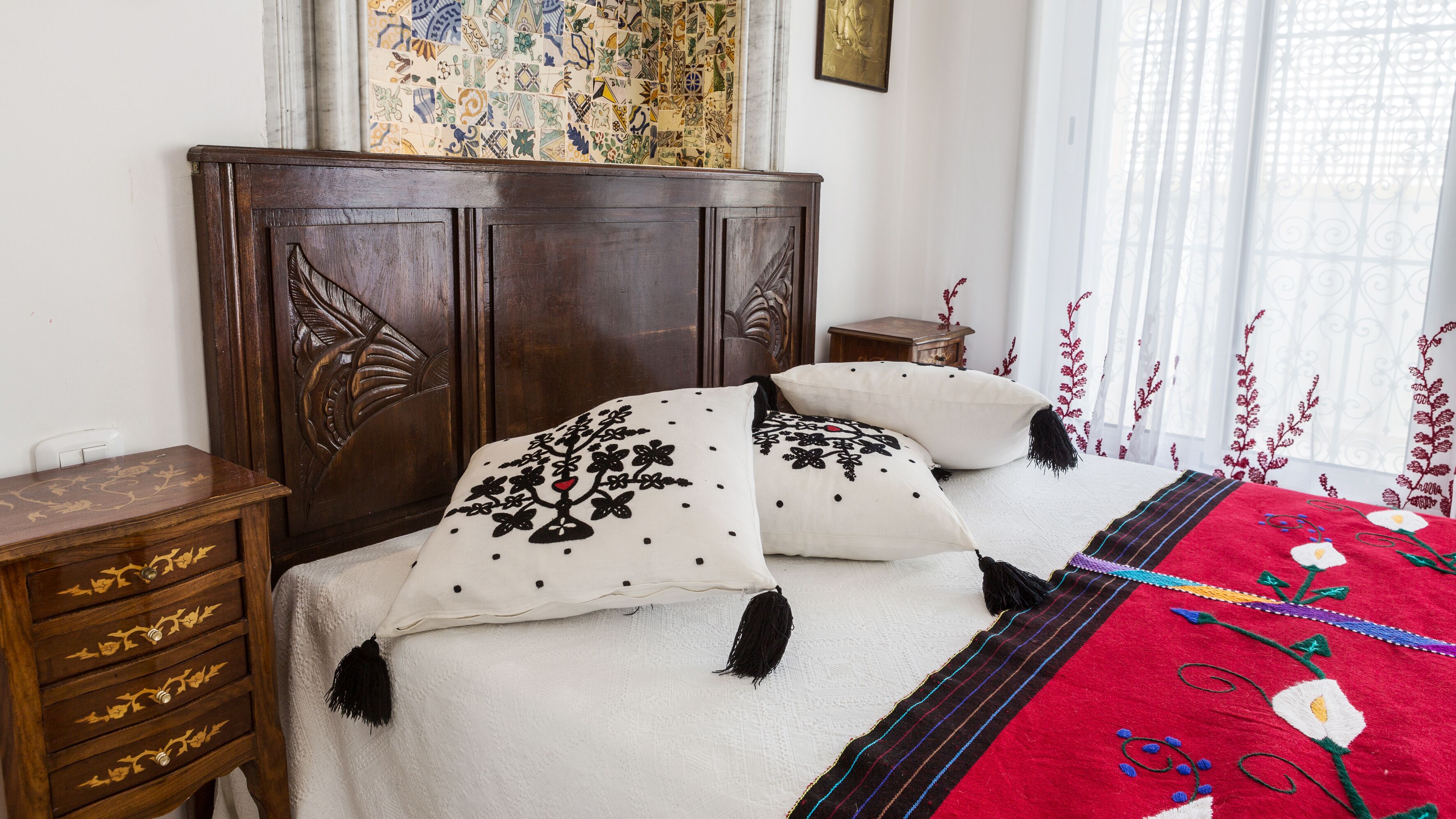 Quarto com cabeceira de madeira, duas camas brancas com almofadas florais pretas, manta vermelha e azul, mesa de cabeceira de madeira e tapeçaria estampada na parede.