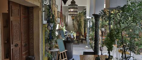 An indoor courtyard with a wooden table and chairs, potted plants, hanging lanterns, and a chalkboard menu stand.